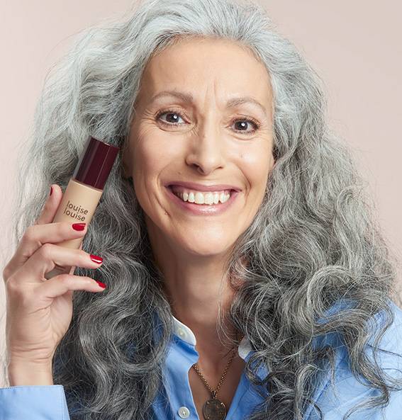 Une très belle femme mature avec un grans sourire tinet dans sa main un flacon du fond de teint Lumière" de la gamme de produits maquillage louiselouise